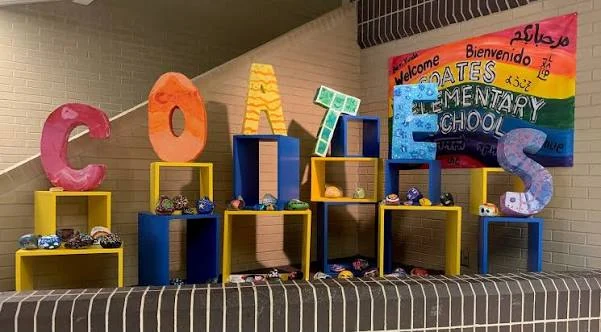 The letters C, O, A, T, E, S in bright colors on stacked shelves of painted rocks, under a rainbow welcome banner for Coates Elementary School in several languages