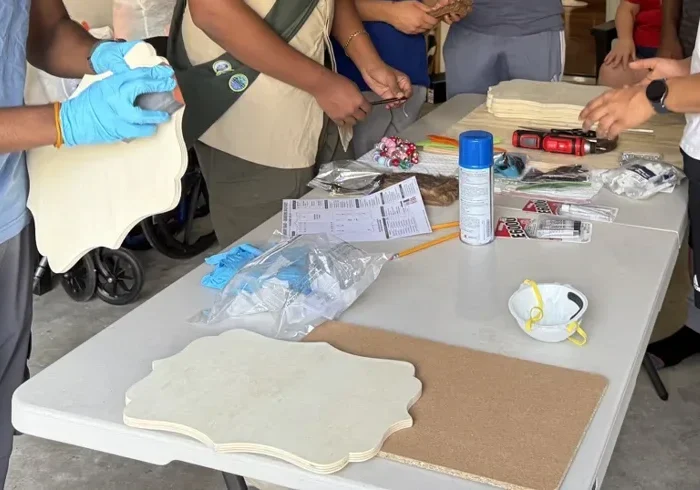 Gloved hands holding a blank board at a worktable covered with hardware, tools and a dust mask
